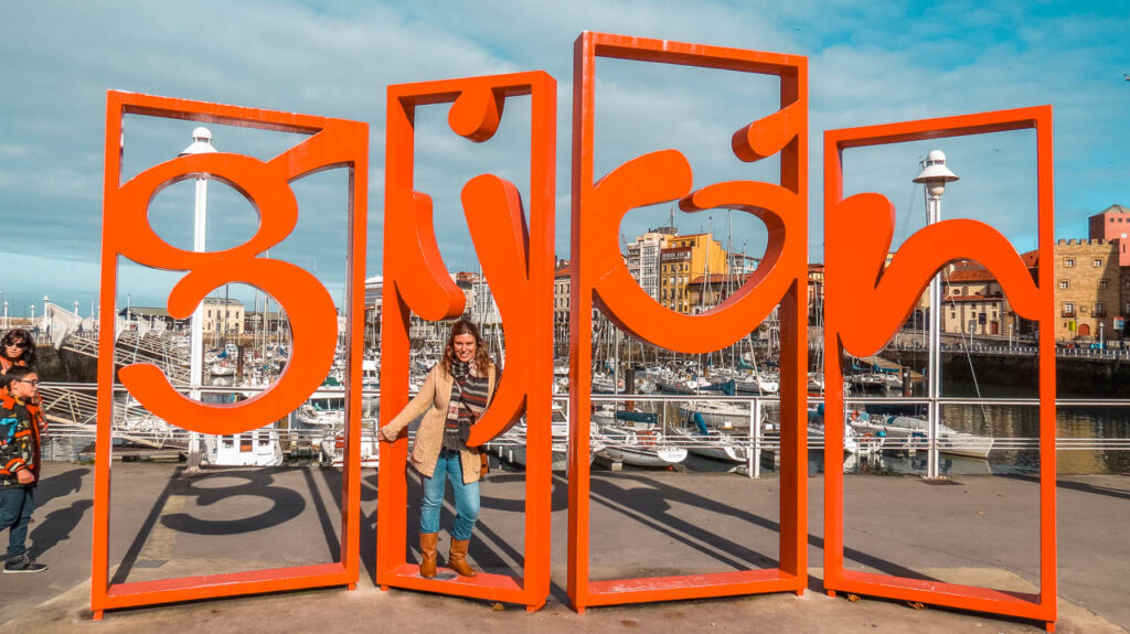 Flor con el cartel de Gijón y de fondo el puerto deportivo